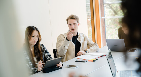 Drei Personen sitzen an einem Tisch, und ein Mann dieser Gruppe zeigt mit seinem Stift in die Richtung der Kamera, wo sich ein Whiteboard befindet. | © FH Kufstein Tirol