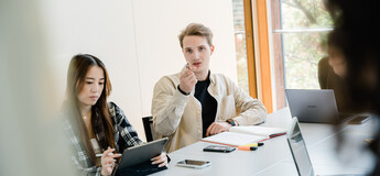 Drei Personen sitzen an einem Tisch, und ein Mann dieser Gruppe zeigt mit seinem Stift in die Richtung der Kamera, wo sich ein Whiteboard befindet. | © FH Kufstein Tirol